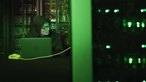 Asian male hacker in hoodie using laptop by computer servers