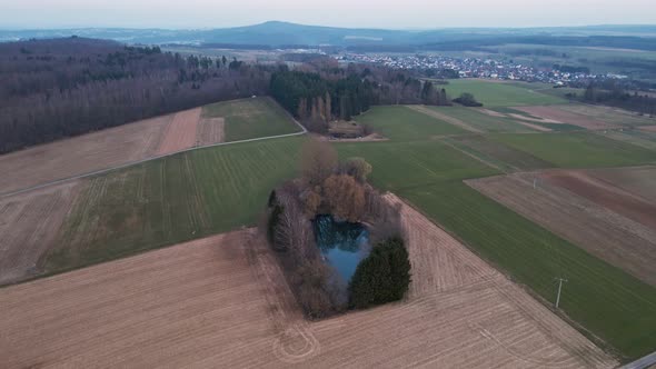 A small lake within green and brown fields in Hesse, Germany. Aerial approach with gimbal tilting do