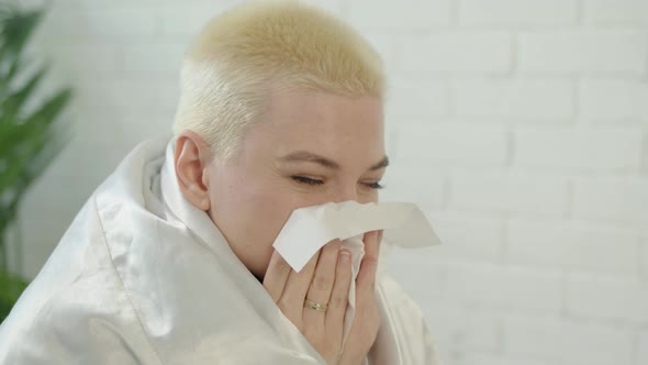 Upset Woman with Short White Hair Wrapped in White Blanket at Home Blows Her Runny Nose Into