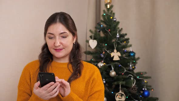 Beautiful Woman with Phone on Background of Christmas Tree