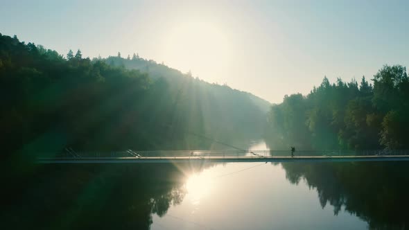 Hiker crossing suspension bridge over river at sunset, dramatic aerial shot