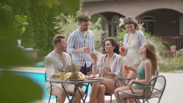 Group of happy young people cheering with cider by the pool in the garden