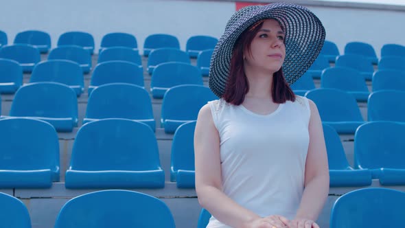 Young Woman in in White Dress and Elegant Hat Looks Away Sitting on Stadium Bleachers Alone