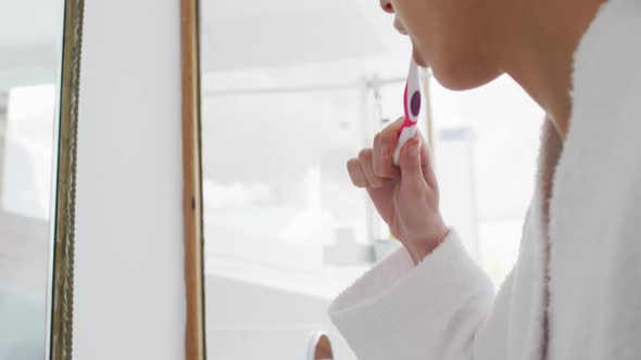 Woman in bathrobe brushing teeth while looking in the mirror