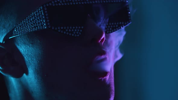 Young Attractive Man in Shades with Rhinestones Vaping in the Dark Club