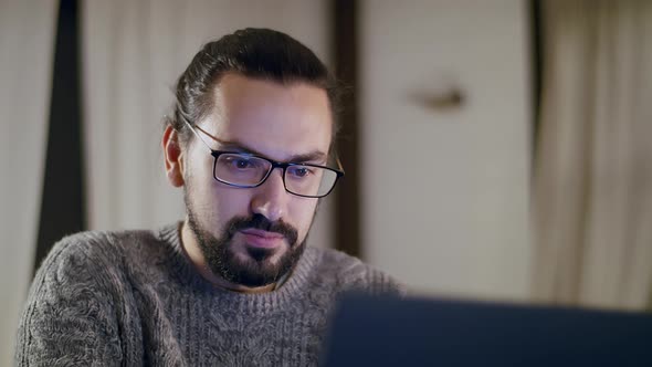 A Young Handsome Caucasian Man in a Knitted Sweater Works at Home Using Laptop