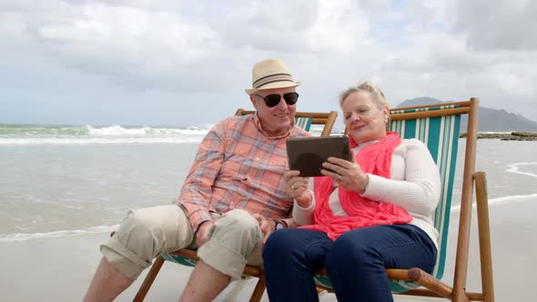Front view of old caucasian senior couple using digital tablet and sitting on sunlounger a beach 4k