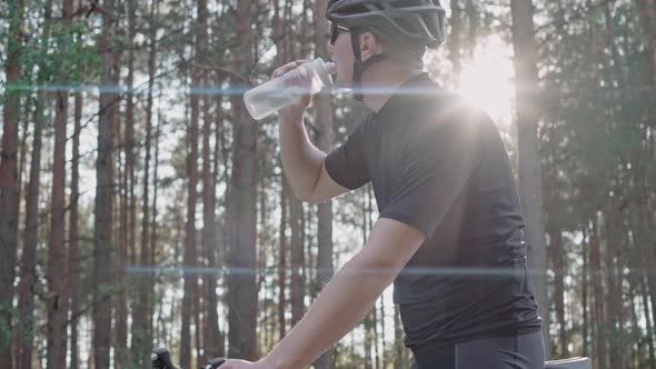 Man Takes Bottle and Drinks Water After Riding on Bike in a Pine Sunny Forest