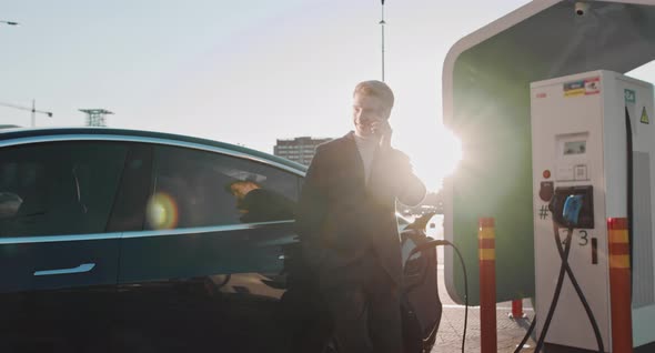 Caucasian Businessman Using Smartphone And Leans On Blue Luxurious Electric Car