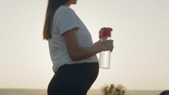 Pregnant Woman Stands in Open Air Against Background of Sun and Sky and Drinks Water From Bottle