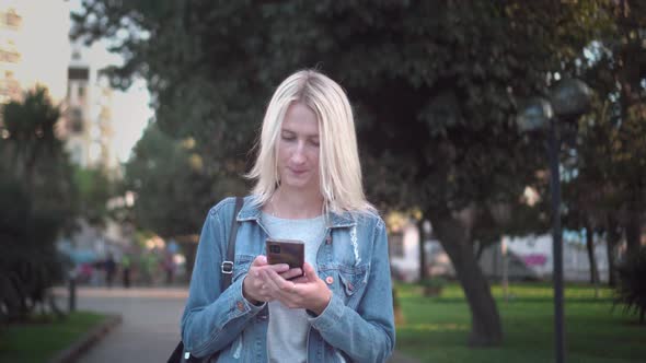 a blonde in a denim jacket walks around the city looking at her smartphone