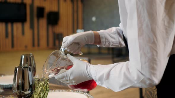 Close Up Waiter Hands in White Gloves Pouring Red Drink From Jug Into Glass Goblet in Restaurant