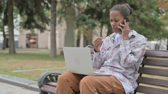 Angry African Woman Talking on Phone and Using Laptop While Sitting Outdoor on Bench