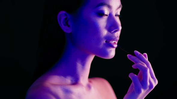 Sensual Asian Woman Applying Lipstick With Brush Standing Under Bright Neon Light