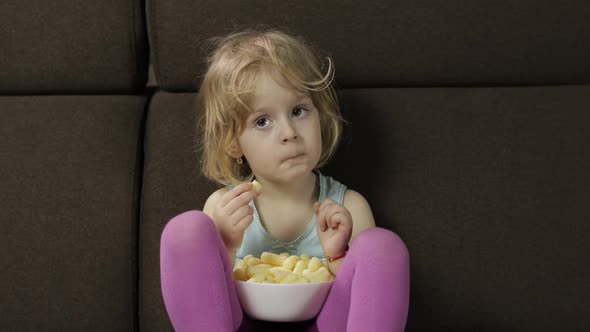 Girl Sitting on Sofa and Eating Corn Puffs. Child Watching Tv, Taste Puffcorns