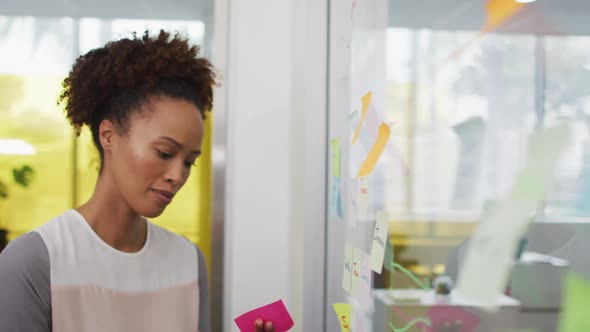 Smiling mixed race businesswoman brainstorming, sticking memo notes on transparent board