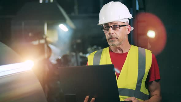 Factory Employee in a Hardhat Is Working with a Laptop