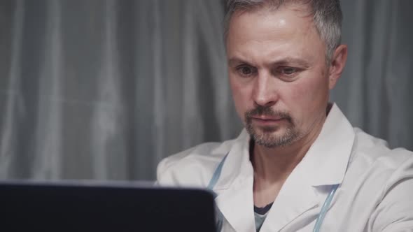 Portrait of Male Doctor Working with Laptop Sitting at Table in Medical Office.