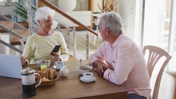 Senior caucasian couple talking to each other using smartphone and laptop having breakfast together