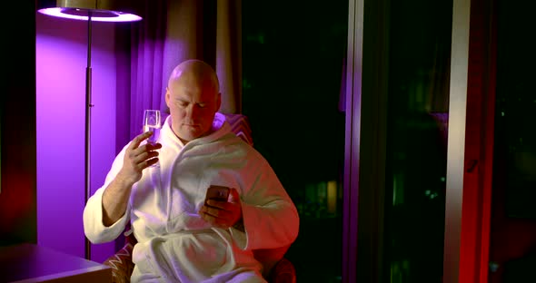 Adult Bald Man, Resting in a Chair After a Shower in a Bathrobe and Writing a Message on a Mobile