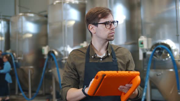 Young Attractive Male Worker Using Digital Tablet and Standing Near Large Metal Tank at Beer Factory