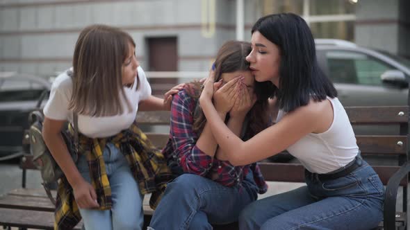 Frustrated Teenage Girl Crying As Friends Calming Down Sitting on Bench on City Street