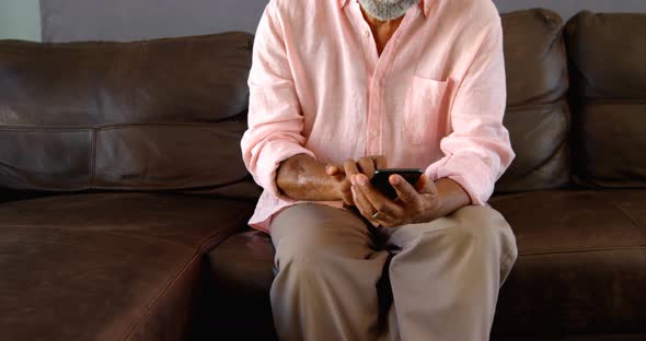 Front view of senior black man sitting on he couch and using mobile phone in a comfortable home 4k