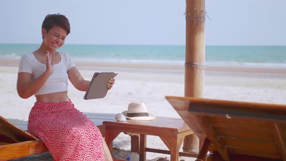 Asian woman chatting with family on video call while relaxing on beach.