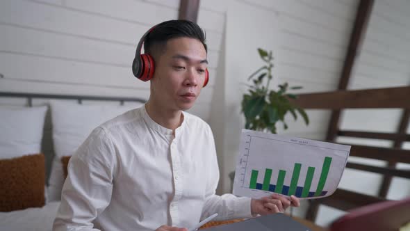 Young Positive Asian Man in Headphones Waving at Web Camera Sitting with Graph in Home Office