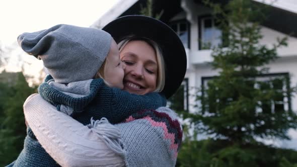 Close-up of a Young Mother Who Holds a Little Boy in Her Arms, Her Son Hugs Tightly Near the House