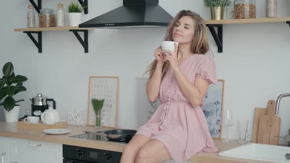 Cute Carefree Girl Enjoying Morning Coffee and Smiling While Sitting on a Table in the Kitchen