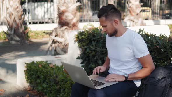 Smiling Young Man Working on the Laptop Sitting in Bench with Travel Case and Bag Backpack Tourist