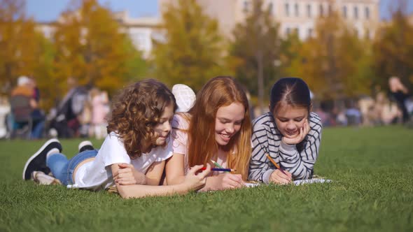 Cute Schoolgirl Friends Relaxing on Green Grass and Drawing with Colorful Pencils