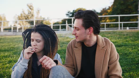 Happy Young Hipster Couple Sitting on Grass and Smoking Marijuana Joint in Park Slow Motion