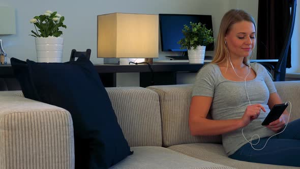 A Young, Beautiful Woman Sits on a Couch in a Living Room and Listens To Music on a Smartphone