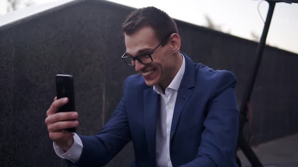 Excited Smiling Businessman Sitting on the Stairs and Does Video Cahtting on Smartphone Outside