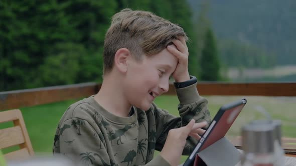 Cute exited boy plays with tablet while sitting outdoor.