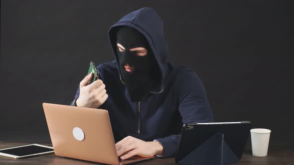 Male Hacker in Black Mask and Hood Uses Laptop To Hack a Hard Drive.