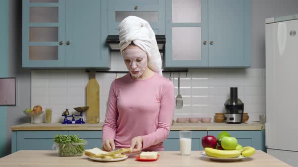 Young Woman in Cloth Face Mask and Towel on Head Eats Sandwich with Butter and Drinks Milk