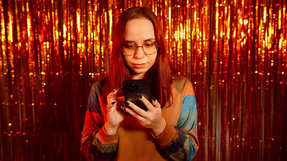 Young Woman in Glasses Browsing Mobile Phone on Background of Shiny Tinsel