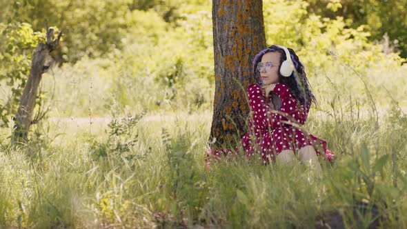 Young Relaxed Woman in Glasses Headphones Sitting Near Tree Listening to Music Dancing Outdoors in