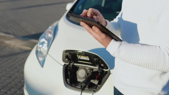 A Lightskinned Male Driver Stands with a Tablet in His Hands Near an Electric Car While It is