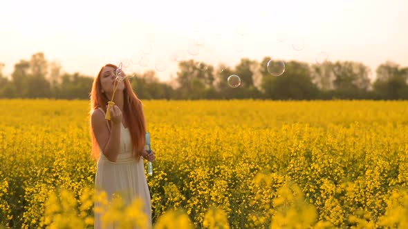 Young Redhair Woman Blowing Bubbles at the Camera Outdoors in Summer Meadow