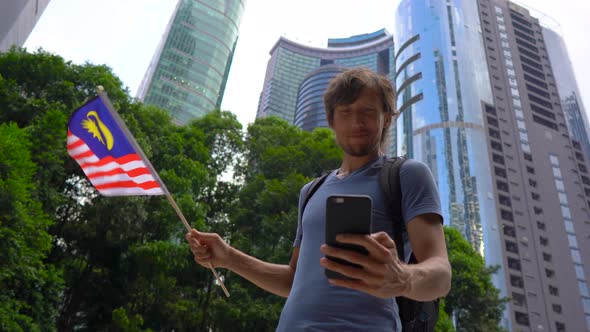 A Young Man Holds a Smartphone and Waves Malaysian Flag with Skyscrapers at a Background