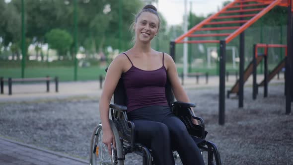 Young Positive Woman in Wheelchair Smiling Looking at Camera Sitting on Sports Ground in Park