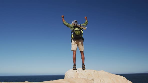 African american man hiking in mountains standing on rock raising his hands by the sea