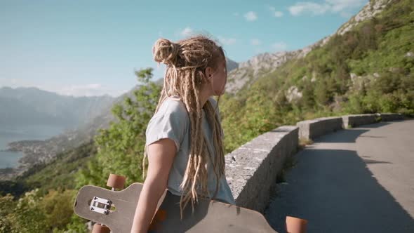 Portrait of  pretty Young dreadlocks woman with longboard standing on mountain background