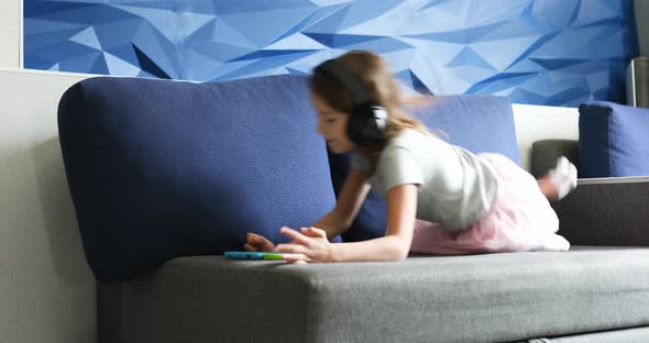 Happy little girl in headphones jumpingon couch with digital tablet, play game