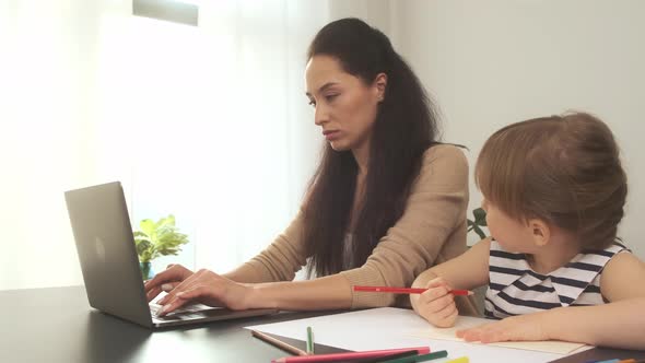 Four Year Old Daughter Draws with Pencils While Her Mother Works Using Laptop in Home Office