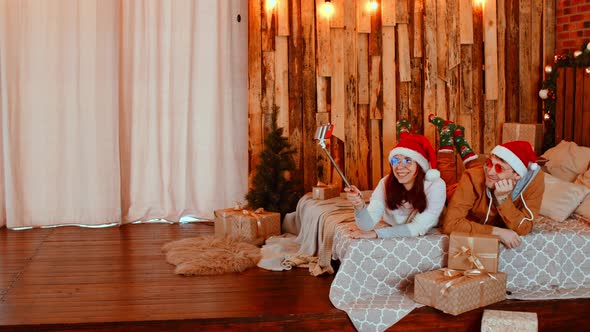 Positive Young Couple in Santa Hats Lying on Comfortable Bed and Taking Selfie on Smartphone